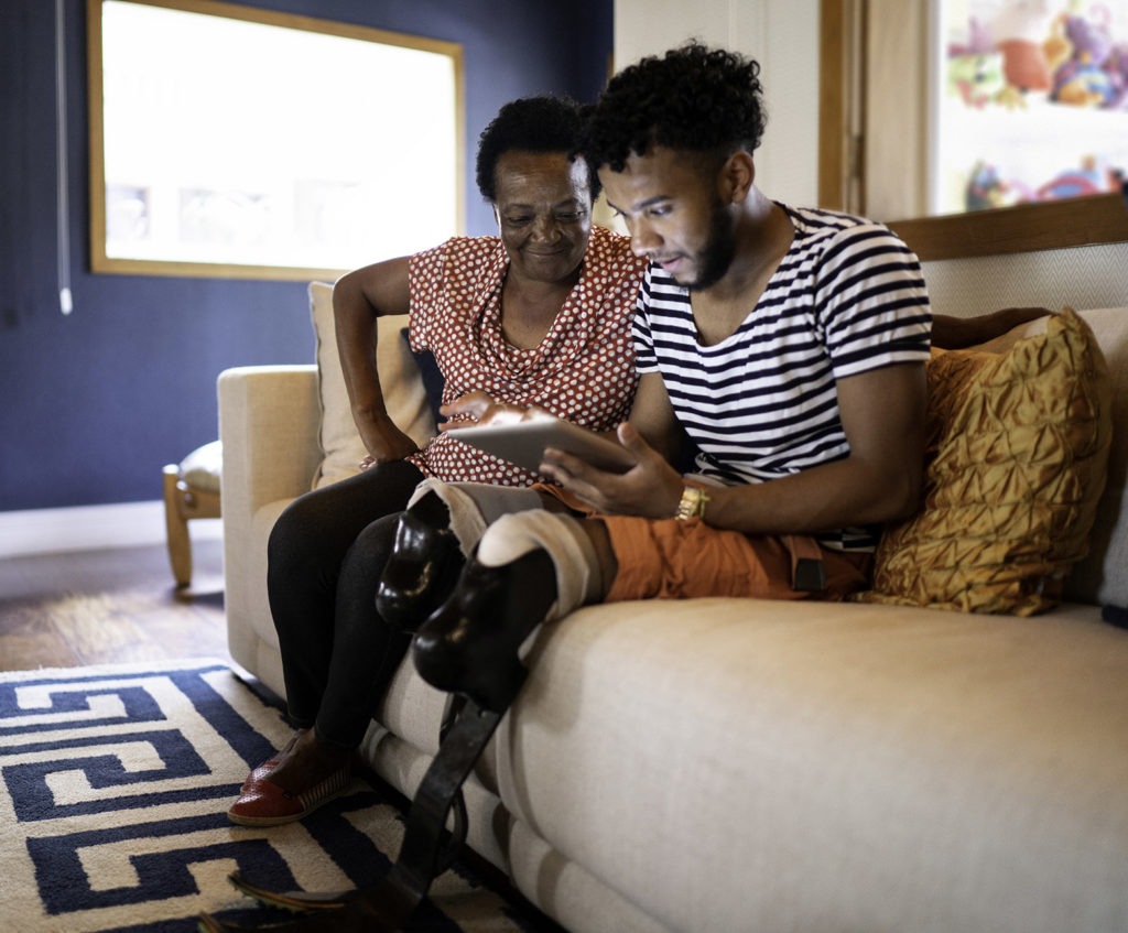 Person with prosthetic leg sits on couch with loved one, both appear upbeat while looking at digital tablet